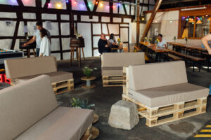 Erste Besucher im Hintergrund an Bierbänlen. Im Vordergrund Europaletten mit Sitzpolstern, die eine entspannte Atmosphäre erzeugen.